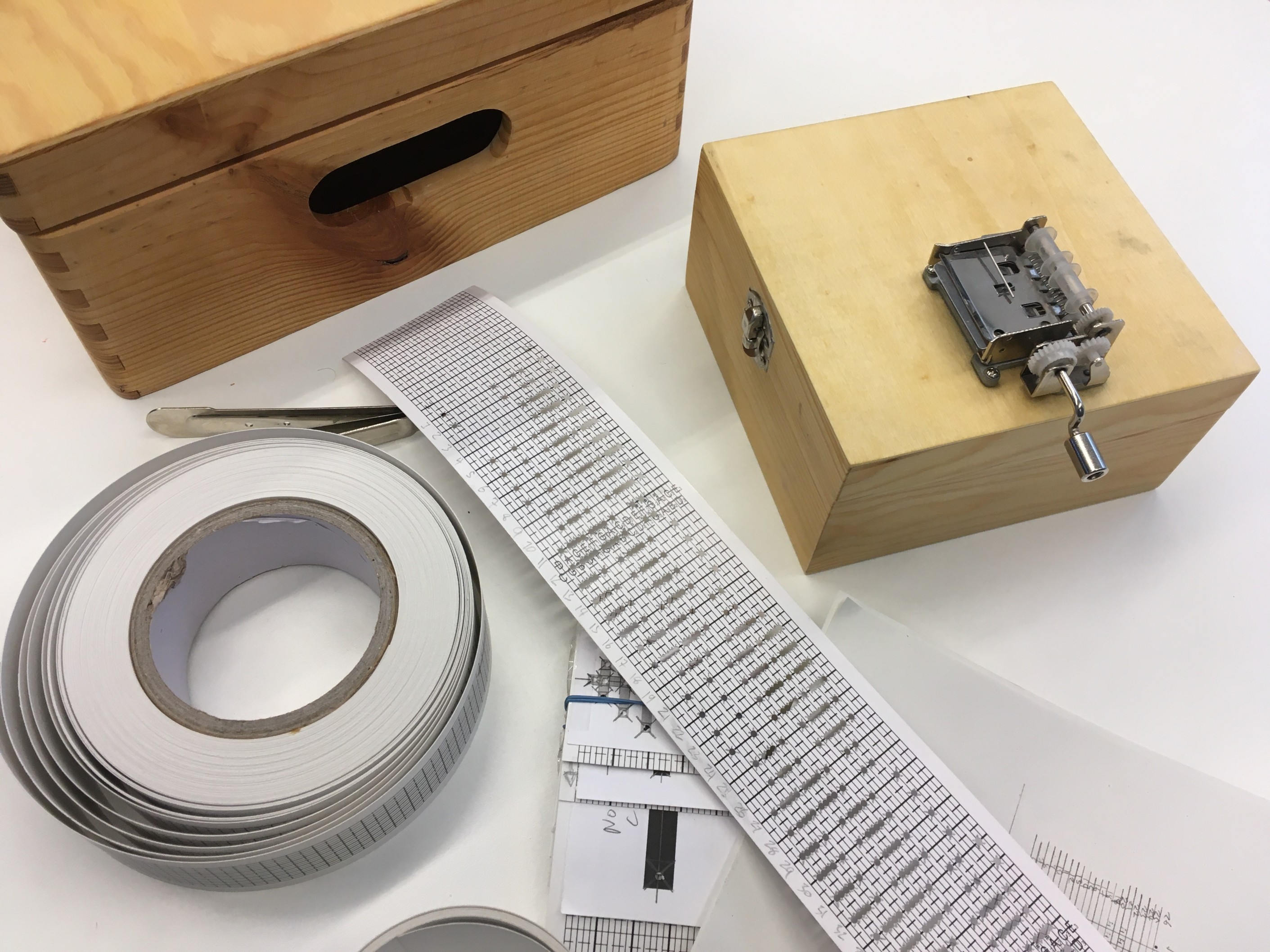 a wooden box and a music box, alongside a punched roll with music values for the box, and an empty roll waiting to be punched with music notes